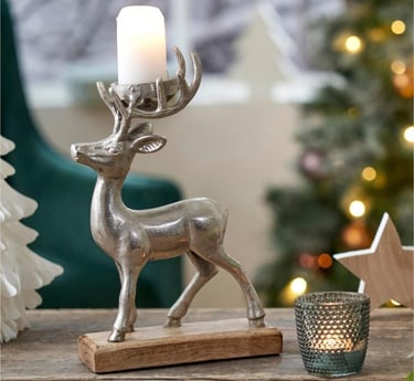 Silver reindeer candle holder on a wooden base with a lit pillar candle near a Christmas tree.